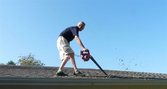 Man on roof using leaf blower to clean gutters.